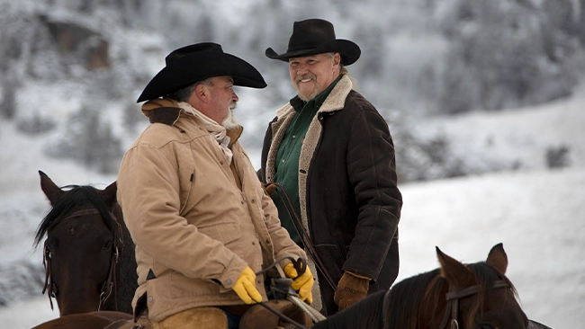 Trace Adkins, host of Ultimate Cowboy Showdown, and another cowboy on horse back having a good laugh together with a snow covered background.